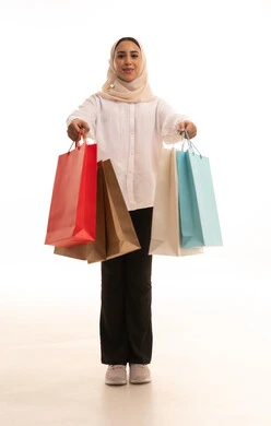 Expressions of happiness and the feeling of enjoyment from shopping, buying gifts and necessities, the positive impact of shopping on women's psychology, benefiting from seasonal offers and discounts, a portrait of a young Arab Gulf Saudi woman wearing a hijab in casual attire holding colorful shopping bags in her hand, full-body shot, white background.