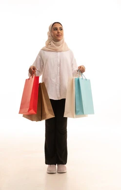Buying gifts and supplies, the positive effect of shopping on women's psychology, gestures of happiness and the feeling of enjoyment in shopping, benefiting from seasonal offers and discounts, a portrait of a young Arab Gulf Saudi woman wearing a hijab in casual attire holding colorful shopping bags in her hand, full-body shot, white background.