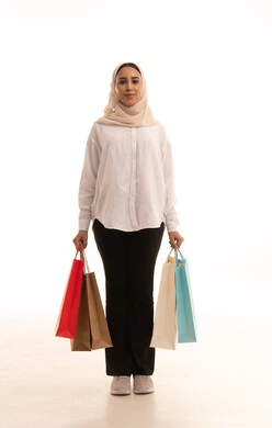 الاُثر الايجابي للتسوق على نفسية النساء، شراء الهدايا والمستلزمات، ايماءات السعادة والشعور بمتعة التسوق، الاستفادة من العروض والتخفيضات الموسميه، بورتريه لشابة عربيه خليجية سعودية محجبة ترتدي الزي الكاجوال  تحمل بيدها أكياس التسوق الملونه, الجسم بالطول الكامل، خلفية بيضاء
