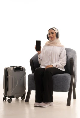 A blank black screen displayed on a mobile phone, using advanced modern technology and devices, a portrait of a young Arab Gulf Saudi woman wearing a hijab in casual attire sitting on a comfortable chair next to a travel bag, wearing headphones and holding a mobile phone with gestures of happiness and joy, white background.