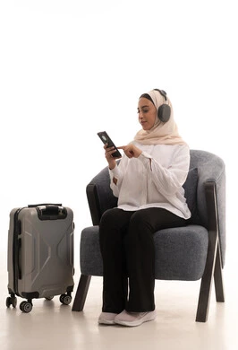 استخدام التكنولوجيا و الأجهزة التقنية الحديثة المتطورة، التسلية والترفيه وقضاء أوقات ممتعه, بورتريه لشابة عربية خليجية سعودية محجبة ترتدي الزي الكاجوال تجلس على كرسي مريح بجانب حقيبة السفر تضع سماعة الهيدفون و تتصفح الهاتف المحمول بايماءات السعادة والسرور, خلفيةبيضاء