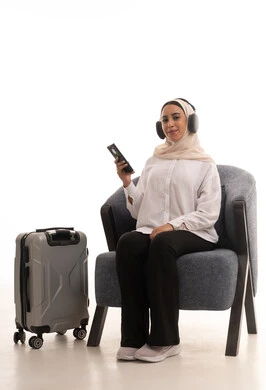 التسلية والترفيه وقضاء أوقات ممتعه, استخدام التكنولوجيا و الأجهزة التقنية الحديثة المتطورة, بورتريه لشابة عربية خليجية سعودية محجبة ترتدي الزي الكاجوال تجلس على كرسي مريح بجانب حقيبة السفر تضع سماعة الهيدفون و تتصفح الهاتف المحمول بايماءات السعادة والسرور, خلفيةبيضاء