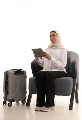 دمج التكنولوجيا المتطورة في الحياة اليومية،  استخدام الاجهزة التقنية الحديثة، الدردشة والتواصل مع الاهل والاصدقاء، بورتريه لشابة عربية خليجية سعودية محجبة ترتدي الزي الكاجوال تجلس على كرسي مريح بجانب حقيبة السفر تحمل بيدها جهاز التابلت، خلفية بيضاء