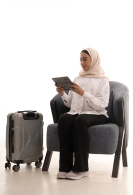 استخدام الاجهزة التقنية الحديثة، الدردشة والتواصل مع الاهل والاصدقاء، دمج التكنولوجيا المتطورة في الحياة اليومية، بورتريه لشابة عربية خليجية سعودية محجبة ترتدي الزي الكاجوال تجلس على كرسي مريح بجانب حقيبة السفر تحمل بيدها جهاز التابلت، خلفية بيضاء