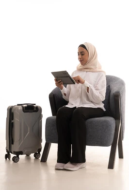 Chatting and communicating with family and friends, using modern technological devices, integrating advanced technology into daily life, a portrait of a young Arab Gulf Saudi woman wearing a hijab in casual attire sitting on a comfortable chair next to a travel bag holding a tablet in her hand, white background.