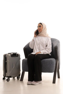 Gestures of focus in listening, communication with family and friends, a portrait of a young Arab Gulf Saudi woman wearing a hijab in casual attire sitting on a comfortable chair holding a mobile phone with a travel bag beside her, making a phone call, using devices, technologies, and travel applications, white background.