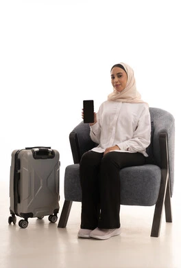 استخدام الأجهزة الحديثة و المتطورة , التجهيز للسفر و الرحلات الممتعة , بورتريه لشابة عربية خليجية سعودية محجبة ترتدي الزي الكاجوال تجلس على كرسي مريح بجانب حقيبة السفر تحمل الهاتف المحمول بشاشة سوداء فارغة , خلفية بيضاء