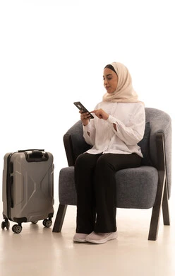 مفهوم السياحة والسفر, بورتريه لشابة عربية خليجية سعودية محجبة ترتدي الزي الكاجوال تجلس على كرسي مريح بجانب حقيبة السفرتمسك بيدها الهاتف المحمول, استخدام التقنيات والتطبيقات السياحية والحجوزات الفندقية, التواصل مع الأصدقاء عبر السوشال ميديا, خلفية بيضاء