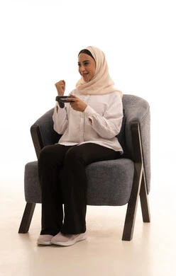 The concept of integration and interaction, electronic games and modern devices, the digital entertainment world, playing and having fun with video games, a portrait of a young Arab Gulf Saudi woman wearing a hijab in casual attire sitting on a comfortable chair holding a game controller, raising her fist in victory gestures, enjoying an exciting entertainment experience, white background.