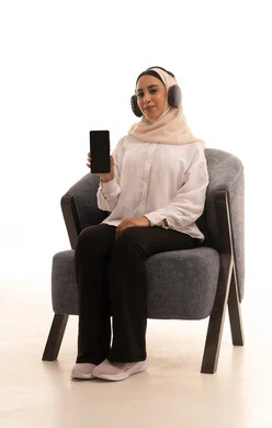 استخدام التكنولوجيا و الأجهزة التقنية الحديثة المتطورة، النظر إلى الكاميرا و الاشارة الى شاشه سوداء فارغه عبر الهاتف المحمول، بورتريه لشابة عربية خليجية سعودية محجبة ترتدي الزي الكاجوال تجلس على كرسي مريح تضع سماعات الرأس، خلفية بيضاء