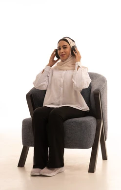 إستخدام تقنيات الهيدفون، مفهوم الإصغاء والإستماع، الاستمتاع في أوقات الفراغ، بورتريه لشابة عربية خليجية سعودية محجبة ترتدي الزي الكاجوال تجلس على كرسي مريح تضع سماعات الرأس الصوتية تستمع إلى الموسيقى، إيماءات السعادة والاندماج، خلفية بيضاء