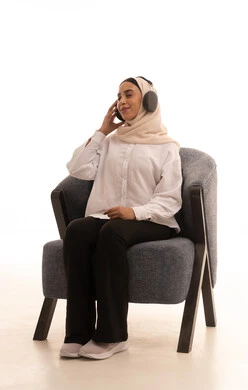 الاستمتاع في أوقات الفراغ, مفهوم الإصغاء والإستماع، إستخدام تقنيات الهيدفون، بورتريه لشابة عربية خليجية سعودية محجبة ترتدي الزي الكاجوال تجلس على كرسي مريحتضع سماعات الرأس الصوتية تستمع إلى الموسيقى، إيماءات السعادة والاندماج، خلفية بيضاء