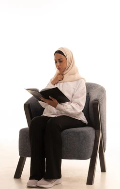 Gestures of engagement and focus, the culture of self-learning, enjoying reading books, stories, and novels, practicing the hobby of reading, spending free time learning, a portrait of a young Arab Gulf Saudi woman wearing a hijab in casual attire sitting on a comfortable chair holding a book in her hand, white background.
