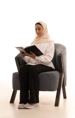 Enjoying reading books, stories, and novels, the culture of self-learning, practicing the hobby of reading, spending free time learning, a portrait of a young Arab Gulf Saudi woman wearing a hijab in casual attire sitting on a comfortable chair holding a book in her hand, white background.