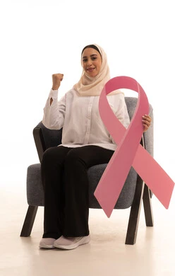 تعابير الانتصار و تحقيق الهدف، حملة توعية للحد من الأمراض الخبيثة, بورتريه لشابة عربية خليجية سعودية محجبة ترتدي الزي الكاجوال تجلس على كرسي تمسك بيدها شعار التوعيه للكشف المبكر عن سرطان الثدي ، خلفية بيضاء