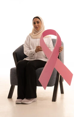 حملة توعية للحد من الأمراض الخبيثة, بورتريه لشابة عربية خليجية سعودية محجبة ترتدي الزي الكاجوال تجلس على كرسي تمسك بيدها شعار التوعيه للكشف المبكر عن سرطان الثدي ، خلفية بيضاء