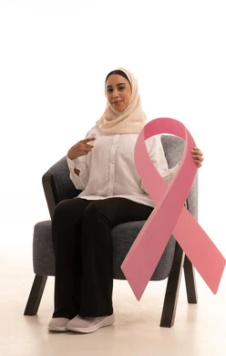 الحملة التوعيه بالامراض الخبيثة, بورتريه لشابة عربية خليجية سعودية محجبة ترتدي الزي الكاجوال تجلس على كرسي تمسك بيدها شعار التوعيه للكشف المبكر عن سرطان الثدي و تشير اليه، خلفية بيضاء