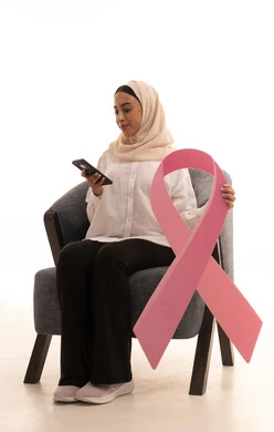 Browsing social media sites via mobile phone, the slogan for World Breast Cancer Awareness Day, the awareness campaign for malignant diseases, a portrait of a young Arab Gulf Saudi woman wearing a hijab in casual attire sitting on a chair holding a pink ribbon, white background.