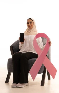 شعار اليوم العالمي للتوعيه بالكشف المبكر لسرطان الثدي، الحملة التوعويه بالامراض الخبيثه، بورتريه لشابة عربية خليجية سعودية محجبة ترتدي الزي الكاجوال تجلس على كرسي تمسك الشريط الوردي، عرض شاشة فارغة سوداء عبر الهاتف المحمول، خلفية بيضاء