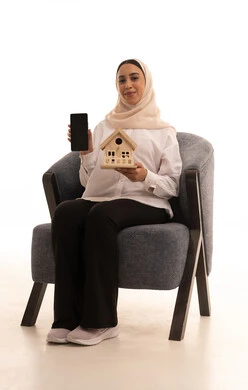 A display of a blank black mobile phone screen, online housing and ownership, buying and selling real estate over the internet, a portrait of a young Arab Gulf Saudi woman wearing casual attire sitting on a chair holding a wooden model of a house with gestures of happiness and joy, white background.