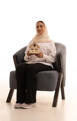 بيع وشراء العقارات، السعي لامتلاك بيت جديد، بورتريه لشابة عربية خليجية سعودية ترتدي الزي الكاجوال تجلس على كرسي وتحمل بيديها مجسم خشبي صغير لمنزل بإيماءات السرور والأمل، خلفية بيضاء