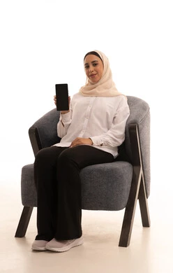 Looking at the camera with gestures of joy and happiness, using advanced modern technology devices, a portrait of a young Arab Gulf Saudi woman wearing a hijab in casual attire sitting on a chair holding a mobile phone with a blank black screen, white background. Looking at the camera with gestures of joy and happiness, using advanced modern technology devices, a portrait of a young Arab Gulf Saudi woman wearing a hijab in casual attire sitting on a chair holding a mobile phone with a blank black screen, white background.