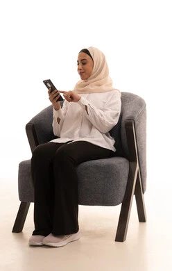 تصفح وسائل التواصل الاجتماعي، الدردشة مع الأهل والأصدقاء، إيماءات السعادة والسرور، استخدام الاجهزة التقنية الحديثة، بورتريه لشابة عربية خليجية سعودية محجبة ترتدي الزي الكاجوال تجلس على كرسي تحمل بيدها الهاتف المحمول، خلفية بيضاء