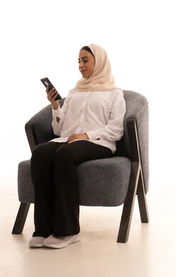 الدردشة مع الأهل والأصدقاء، إيماءات السعادة والسرور، استخدام الاجهزة التقنية الحديثة، بورتريه لشابة عربية خليجية سعودية محجبة ترتدي الزي الكاجوال تجلس على كرسي تحمل بيدها الهاتف المحمول، تصفح وسائل التواصل الاجتماعي، خلفية بيضاء