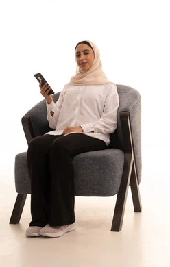 إيماءات السعادة والسرور، الدردشة مع الأهل والأصدقاء، استخدام الاجهزة التقنية الحديثة، بورتريه لشابة عربية خليجية سعودية محجبة ترتدي الزي الكاجوال تجلس على كرسي تحمل بيدها الهاتف المحمول، تصفح وسائل التواصل الاجتماعي، خلفية بيضاء