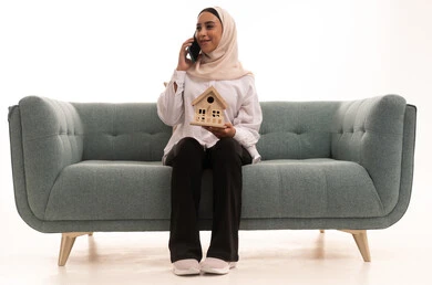 Making a phone call via mobile phone, benefiting from modern technologies in the real estate field, buying and selling properties through mobile, a portrait of a young Arab Gulf Saudi woman wearing casual attire sitting on a couch holding a wooden model of a house in her hands, white background.