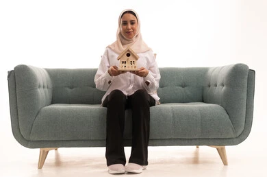 السعي لامتلاك بيت جديد، بيع وشراء العقارات، بورتريه لشابة عربية خليجية سعودية ترتدي الزي الكاجوال تجلس على كنبة مريحة وتحمل بيديها مجسم خشبي صغير لمنزل بإيماءات السرور والأمل، خلفية بيضاء
