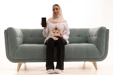 عرض شاشة هاتف محمول سوداء فارغة، الإسكان والتمليك أونلاين، بيع وشراء العقارات عبر الانترنت، بورتريه لشابة عربية خليجية سعودية محجبة ترتدي الزي الكاجوال تجلس على كنبة وتحمل بيديها مجسم خشبي لمنزل بإيماءات السعادة والسرور، خلفية بيضاء