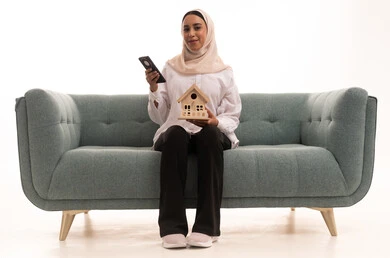 الإسكان والتمليك أونلاين عبر الهاتف المحمول، الاستفادة من التقنيات الحديثة في المجال العقاري، بيع وشراء العقارات عبر الانترنت، بورتريه لشابة عربية خليجية سعودية محجبة ترتدي الزى الكاجوال تجلس على الاريكة وتحمل بيديها مجسم خشبي لمنزل، خلفية بيضاء