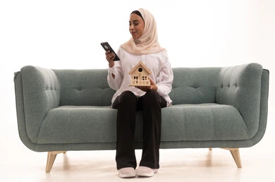 الاستفادة من التقنيات الحديثة في المجال العقاري، الإسكان والتمليك أونلاين عبر الهاتف المحمول، بيع وشراء العقارات عبر الانترنت، بورتريه لشابة عربية خليجية سعودية محجبة ترتدي الزى الكاجوال تجلس على الاريكة وتحمل بيديها مجسم خشبي لمنزل، خلفية بيضاء