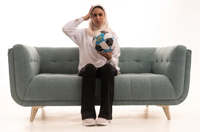 مفهوم الاستياء لعدم فوز الفريق المفضل، قضاء وقت ممتع بمتابعة المباراة، بورتريه لشابة عربية خليجية سعودية محجبة ترتدي الزى الكاجوال تجلس على الاريكة تحمل كرة القدم تضع يديها على رأسه بإيماءات الحزن والخسارة، المباريات والألعاب الرياضية، خلفية بيضاء