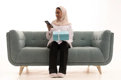 Browsing social media sites via mobile phone, the concept of gifting and appreciation, a portrait of a young Arab Gulf Saudi woman wearing casual attire sitting on a sofa holding a closed box in her hands, acquiring and choosing gifts, white background. Browsing social media sites via mobile phone, the concept of gifting and appreciation, a portrait of a young Arab Gulf Saudi woman wearing casual attire sitting on a sofa holding a closed box in her hands, acquiring and choosing gifts, white background.