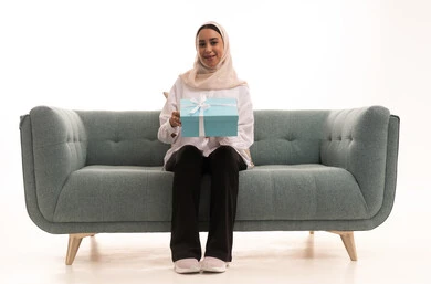 Happy occasions and holidays, gestures of joy at receiving commemorative gifts, the concept of gifting and appreciation, a portrait of a young Arab Gulf Saudi woman wearing a hijab in casual attire sitting on a sofa holding a closed box in her hands, acquiring and choosing gifts, white background.