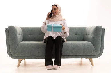 Gift-giving during holidays and occasions to express love, the concept of gifting and appreciation, a portrait of a young Arab Gulf Saudi woman wearing a hijab in casual attire sitting on a sofa holding a gift box in her hand, making a phone call on her mobile, gestures of happiness and joy, white background.