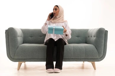 ايماءات المفجاءة و الاندهاش لسماع خبر ما، مفهوم الإهداء والتقدير، تقديم الهدايا في الاعياد والمناسبات للتعبير عن المحبة، بورتريه لشابة عربية خليجية سعودية محجبة ترتدي الزى الكاجوال تجلس على الاريكة تحمل بيدها علبة هدايا، اجراء مكالمة هاتفية عبر الجوال، خلفية بيضاء