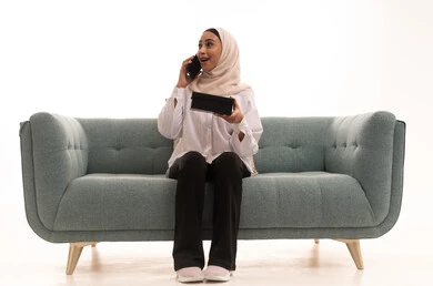 The concept of gifting and appreciation, giving gifts during holidays and occasions to express love, a portrait of a young Arab Gulf Saudi woman wearing a hijab and casual attire sitting on a sofa holding a gift box in her hand, making a phone call on her mobile, gestures of happiness and joy, white background.
