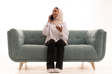 التواصل مع الاهل والاصدقاء عبر الجوال، ايماءات السعادة والسرور، بورتريه لشابة عربية خليجية سعودية محجبة ترتدي الزى الكاجوال تجلس على الاريكة تقوم باجراء مكالمة هاتفية عبر الهاتف المحمول، دمج التقنيات الحديثة بالحياة اليومية، خلفية بيضاء