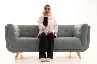عرض تطبيقات الهاتف المحمول، التسويق الالكتروني باستخدام التقنية والتكنولوجيا، بورتريه لشابة عربية خليجية سعودية محجبة ترتدي الزى الكاجوال تجلس على الاريكة تحمل بيدها جوال بشاشة سوداء فارغة، النظر الى الكاميرا بتعابير السرور، خلفية بيضاء