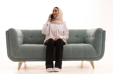 Gestures of happiness and joy, a portrait of a young Arab Gulf Saudi woman wearing a hijab and casual attire sitting on a sofa making a phone call via her mobile phone, integrating modern technologies into daily life, communicating with family and friends through the mobile phone, white background.