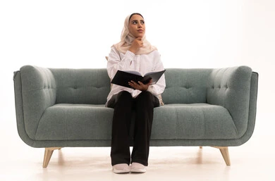 النظر و التفكير فى شيء ما، هواية القراءة والاستكشاف، قضاء وقت الفراغ بالتعلم والثقافة, مفهوم التثقيف والبحث العلمي, بورتريه لشابة عربية خليجية سعودية محجبة ترتدي الزى الكاجوال تجلس على الاريكة تقوم بتصفح المجلدات العلميه والثقافيه، خلفيه بيضاء