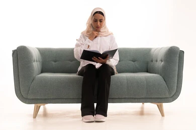 هواية القراءة والاستكشاف، ايماءات الاندماج و التركيز، قضاء وقت الفراغ بالتعلم والثقافة, مفهوم التثقيف والبحث العلمي, بورتريه لشابة عربية خليجية سعودية محجبة ترتدي الزى الكاجوال تجلس على الاريكة تقوم بتصفح المجلدات العلميه والثقافيه، خلفيه بيضاء