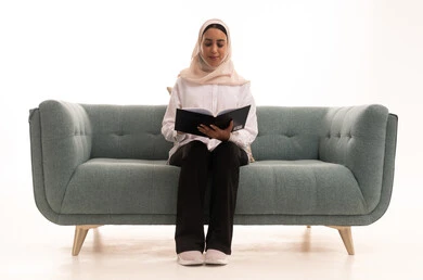 Gestures of integration and focus, spending free time on learning and culture, the hobby of reading and exploration, the concept of education and scientific research, a portrait of a young Arab Gulf Saudi woman wearing a hijab in casual attire sitting on a sofa browsing scientific and cultural volumes, white background.