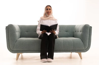 قضاء وقت الفراغ بالتعلم والثقافة, هواية القراءة والاستكشاف, مفهوم التثقيف والبحث العلمي, بورتريه لشابة عربية خليجية سعودية محجبة ترتدي الزى الكاجوال تجلس على الاريكة تقوم بتصفح المجلدات العلميه والثقافيه، خلفيه بيضاء