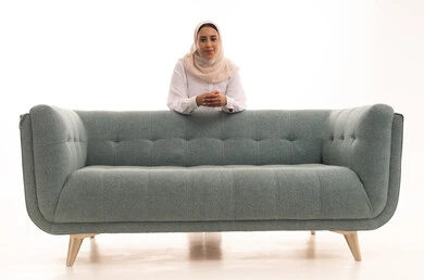 نظرات الأنوثة الناعمة, إيماءات السرور والسعادة، الاهتمام بالمظهر الخارجي, بورتريه لشابة عربية خليجية سعودية محجبة ترتدي الزى الكاجوال تقف بايدي متشابكة خلف الاريكة تنظر إلى الكاميرا, الاستمتاع بقضاء وقت ممتع, خلفية بيضاء