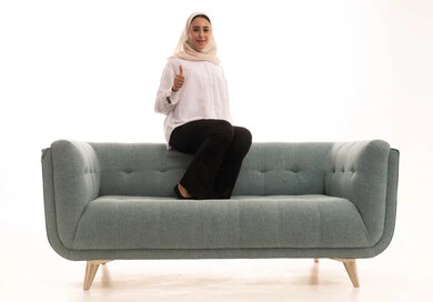 Expressions of acceptance and approval, gestures indicating quality and excellence, gestures of joy and happiness, a portrait of a young Arab Gulf Saudi woman wearing a hijab in casual attire sitting on a sofa pointing her thumb up, enjoying a good time, soft feminine looks, white background.