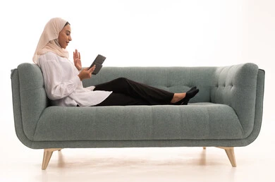 Raising the palm of the hand with expressions of peace and greeting, making a video call over the internet, having a good time on the mobile phone with friends, a portrait of a Saudi Arabian Gulf woman wearing a hijab in casual attire sitting on the couch using a mobile phone, white background.